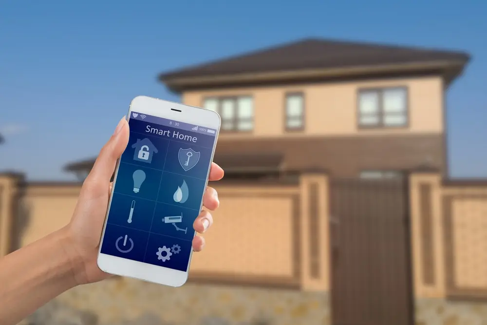 Mão segurando um smartphone exibindo um app para controlar equipamentos de uma casa inteligente ao fundo.