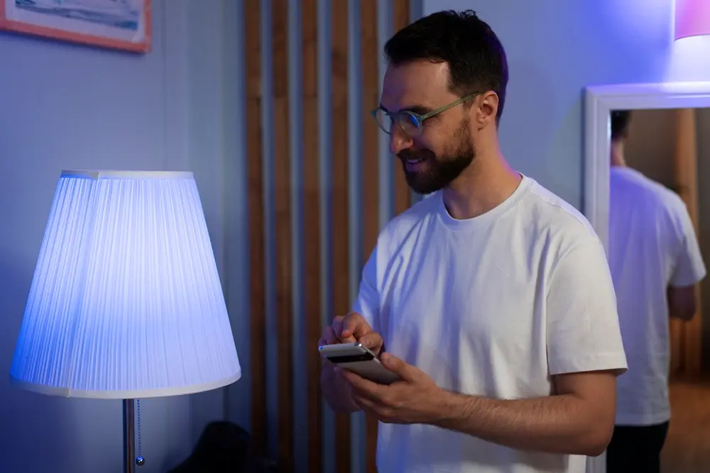 Homem usando uma camisa cor branca com um smartphone na mão controlando a cor de uma lâmpada inteligente.