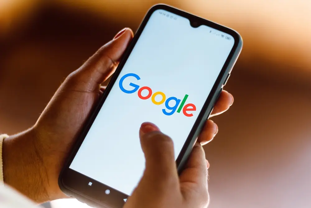 Mãos segurando um smartphone exibindo a tela do buscador Google