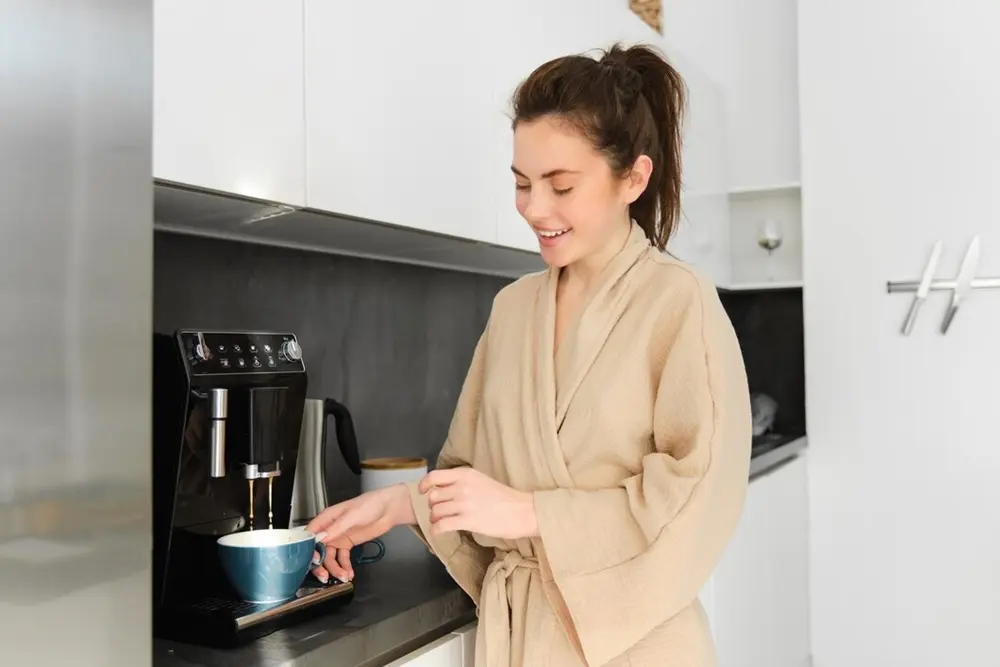 Uma mulher vestindo um roupão cor bege segurando uma xicara em uma cafeteira smart.