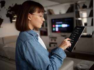 Uma pessoa vestindo uma jaqueta jeans segura um tablet exibindo uma interface de controle de TV inteligente em uma sala de estar moderna com um mapa-múndi na parede.