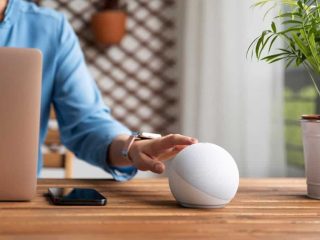 Uma pessoa de camisa azul interage com um alto-falante inteligente branco em uma mesa de madeira, com um laptop e um smartphone por perto. Uma planta está ao fundo.