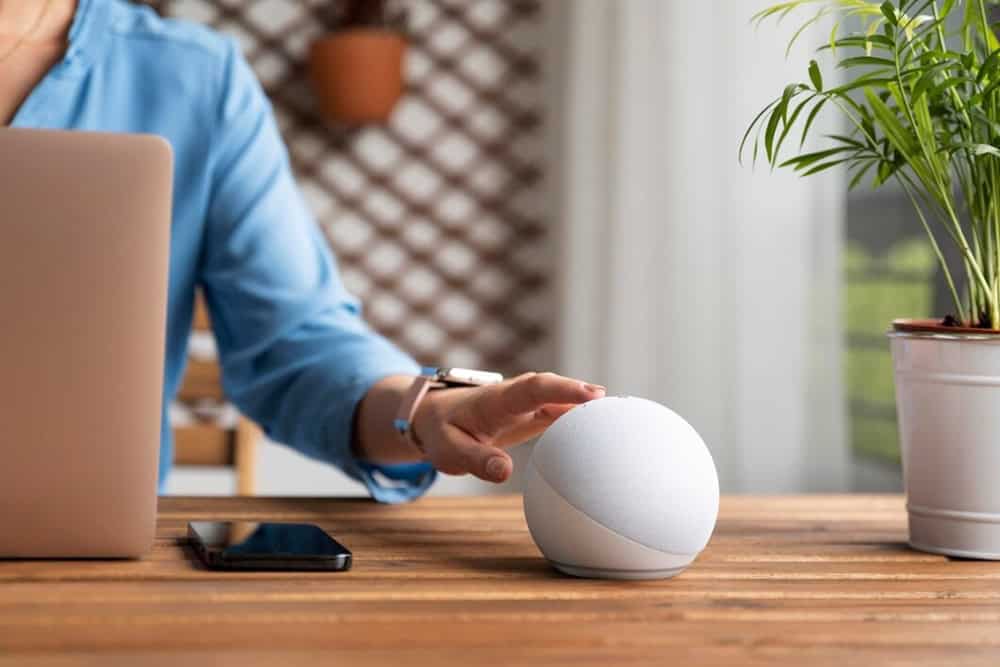 Uma pessoa de camisa azul interage com um alto-falante inteligente branco em uma mesa de madeira, com um laptop e um smartphone por perto. Uma planta está ao fundo.