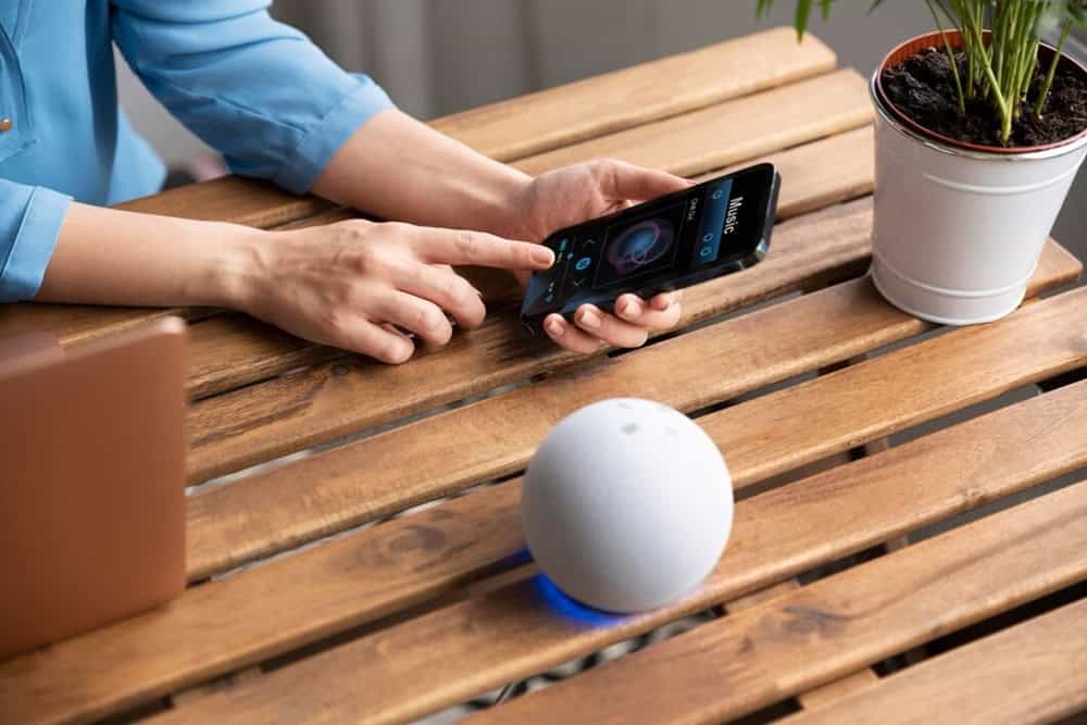 Uma pessoa de camisa azul interage com um alto-falante inteligente branco em uma mesa de madeira, com um laptop e um smartphone por perto. Uma planta está ao fundo. Como Personalizar as Respostas da Alexa às Rotinas da Amazon