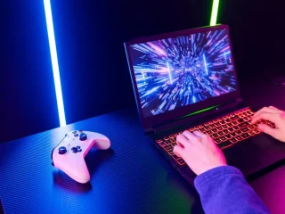 Uma pessoa com as mãos sobe um teclado iluminado de um notebook gamer e um joystick ao lado.