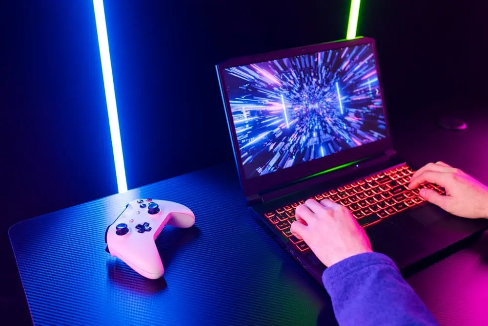 Uma pessoa com as mãos sobe um teclado iluminado de um notebook gamer e um joystick ao lado.