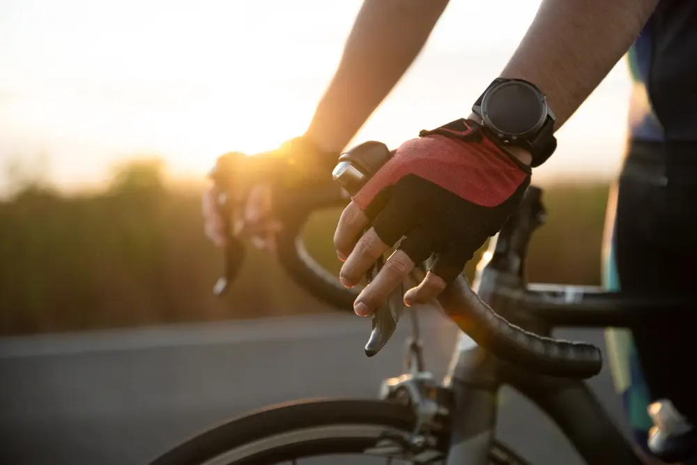 Braço de um homem segurando o guidão de uma bike de estrada usando luvas de ciclismo e um smartwtch. Smartwatch para ciclismo com GPS