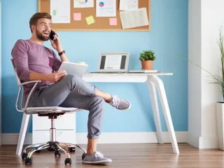Uma pessoa sentada confortavelmente em um home office, segurando um tablet e falando ao telefone, cercada por uma decoração elegante.