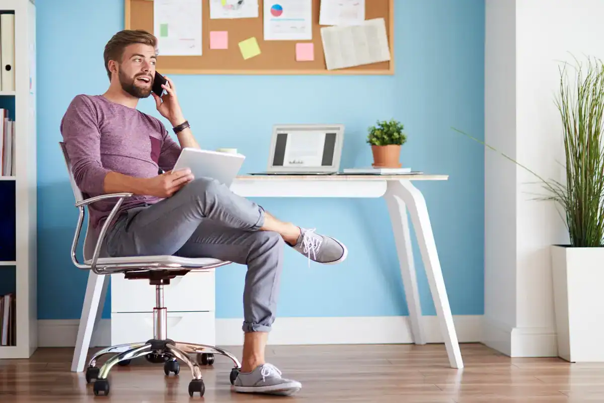 Uma pessoa sentada confortavelmente em um home office, segurando um tablet e falando ao telefone, cercada por uma decoração elegante.