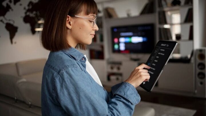 Uma pessoa vestindo uma jaqueta jeans segura um tablet exibindo uma interface de controle de TV inteligente em uma sala de estar moderna com um mapa-múndi na parede.