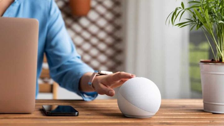 Uma pessoa de camisa azul interage com um alto-falante inteligente branco em uma mesa de madeira, com um laptop e um smartphone por perto. Uma planta está ao fundo.