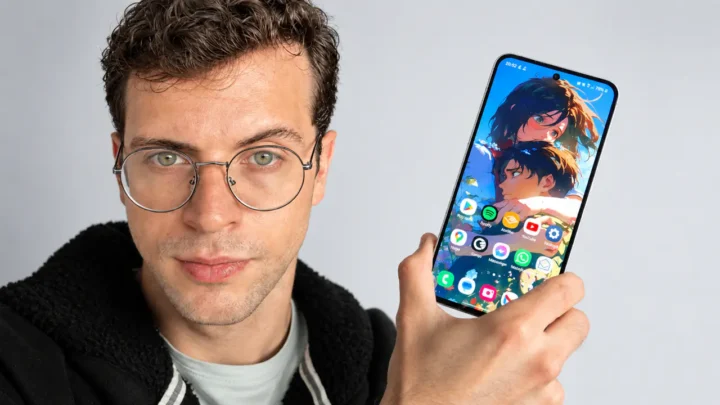 Homem jovem com cabelos castanhos ondulados, óculos redondos e barba rala segura um smartphone Samsung Galaxy S24 FE com tela colorida diante de um fundo cinza-claro, em uma composição ultrarrealista de estilo publicitário tecnológico.